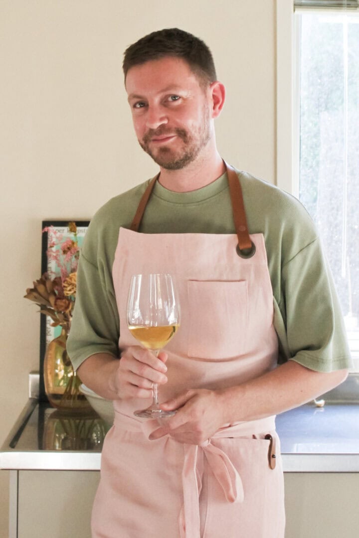 Portrait of Ben Walls in his kitchen wearing a green t-shirt and a pink apron, holding a glass of white wine.