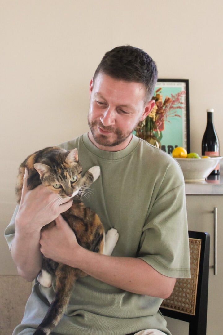 Portrait of Ben Walls in his kitchen wearing a green t-shirt, holding his tortoiseshell tabby cat, Billie.