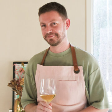 Portrait of Ben Walls in his kitchen wearing a green t-shirt and a pink apron, holding a glass of white wine.