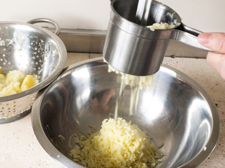 Cooked agria potatoes being passed through a potato ricer into a large mixing bowl.