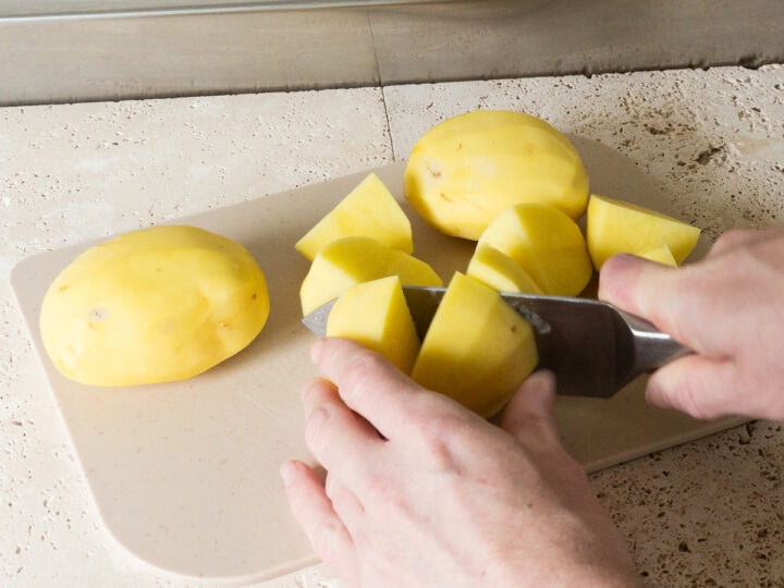 Peeled agria potatoes being cut into quarters on a chopping board.