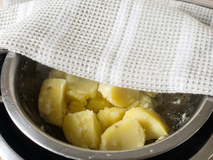 Boiled potato quarters steaming in a colander under a tea towel.