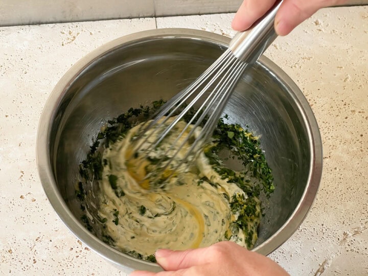 Mixed herbs, garlic, lemon zest and sour cream being mixed into mayonnaise with a whisk.