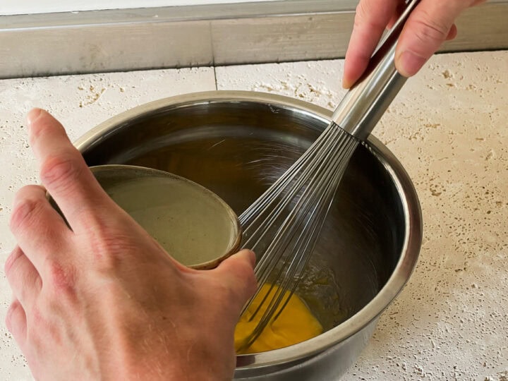 Oil being added slowly into a bowl and emulsified with egg yolk to make mayonnaise using a whisk.