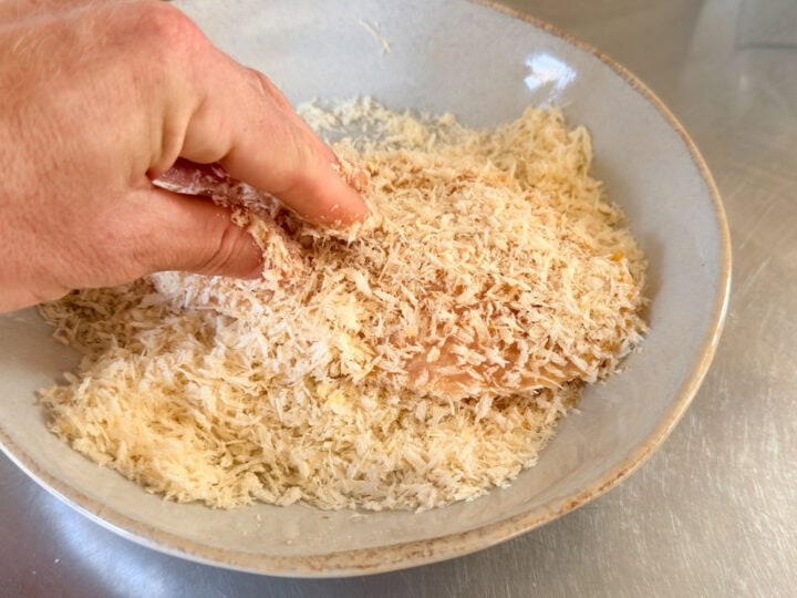 Close-up of hand pressing breadcrumbs onto chicken cutlet in a mixing bowl.