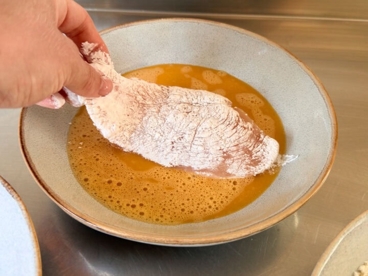 Chicken cutlet dredged in cornflour being dipped in beaten egg.