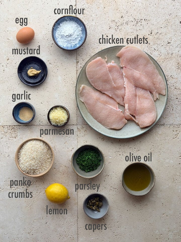 Raw chicken cutlets with ingredients for crumbing to make chicken cotoletta: whole egg, cornflour, mustard, garlic powder, finely grated parmesan, panko crumbs, whole lemon, chopped parsley, capers, olive oil.