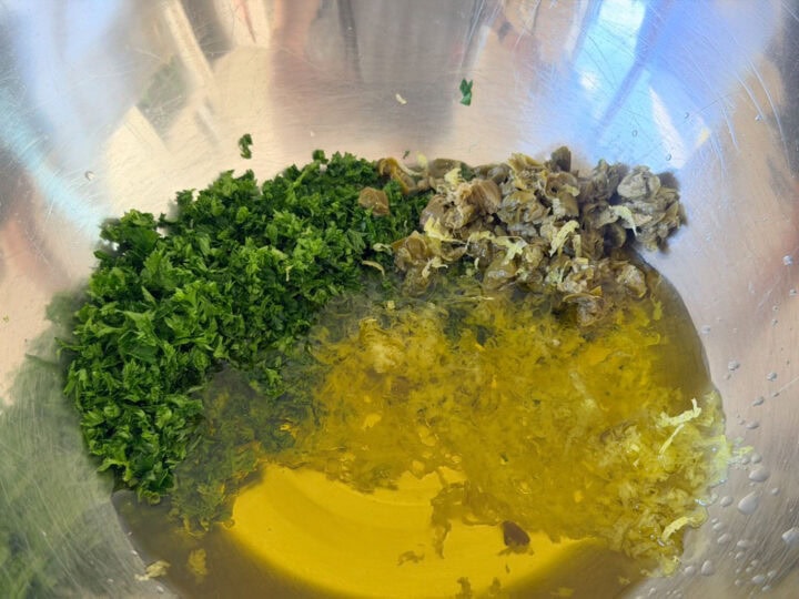 Fresh parsley, capers and lemon zest with olive oil and lemon juice in a stainless steel bowl.
