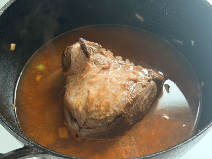 Browned beef cheek in a cast iron pot with cooking liquid and diced vegetables.