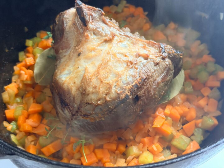 Browned beef cheek sitting on top of sautéed onions, carrot and celery, with thyme and bay leaves in a cast iron pot.