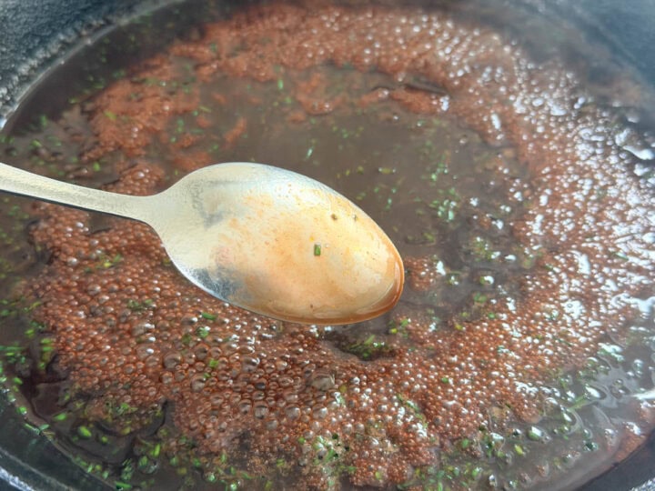 A sauce with finely chopped chives being reduced in a cast iron skillet with a stainless steel spoon that has the sauce coating the back of it.