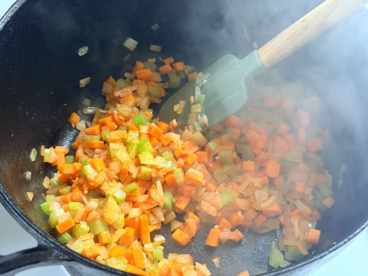 Carrots, onion and celery being sautéed in a cast iron pot and stirred with a green spatula.