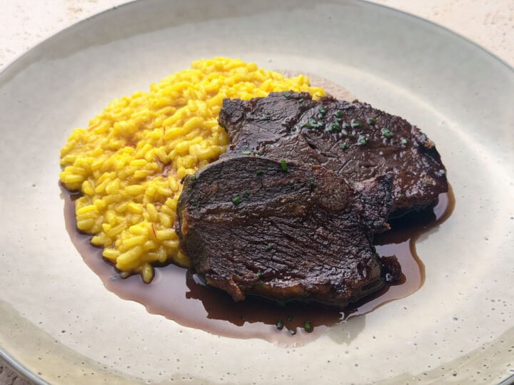 Two slices of tender, red wine braised beef cheeks, with a rich sauce and chopped chives, served with a saffron risotto to the side on a green plate.