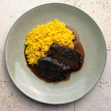 A Birds Eye view of two slices of tender, red wine braised beef cheeks, with a rich sauce and chopped chives, served with a saffron risotto to the side on a green plate.
