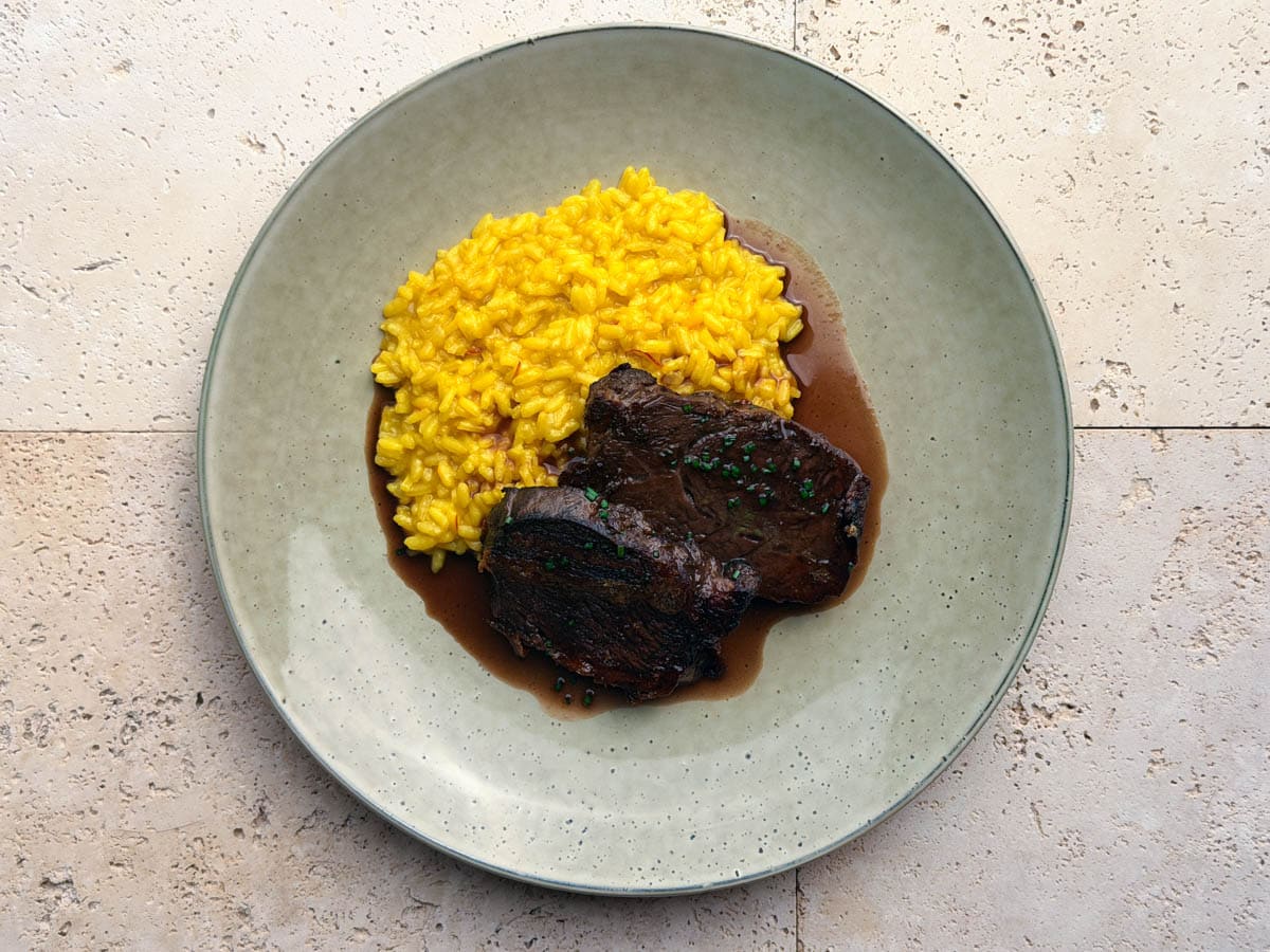A Birds Eye view of two slices of tender, red wine braised beef cheeks, with a rich sauce and chopped chives, served with a saffron risotto to the side on a green plate.