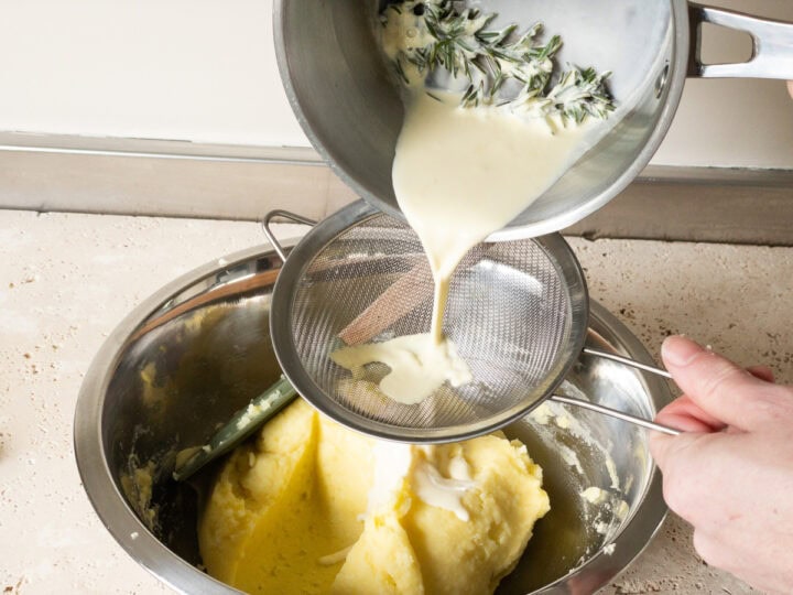 Rosemary infused cream being poured through a sieve into a large mixing bowl of mashed potatoes.